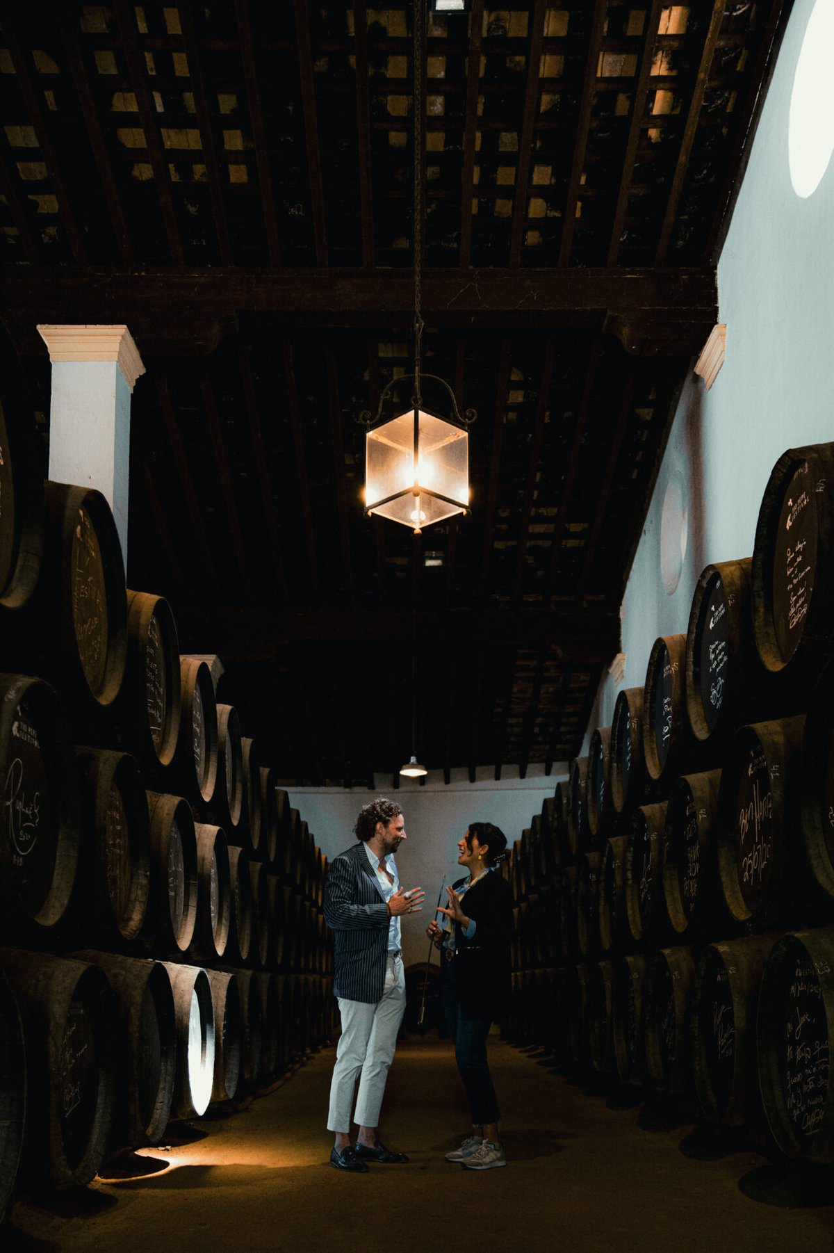 Two people talking between towering rows of dark sherry barrels under a hanging lantern