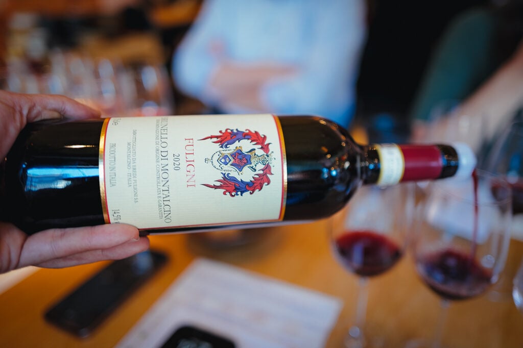 A hand holds a Fuligni Brunello di Montalcino 2020 bottle while red wine is poured behind it