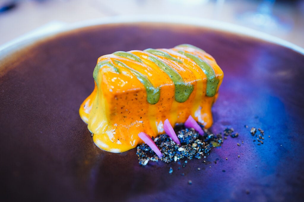 A plated dish in orange and green sauce with pink onion garnish on a dark ceramic plate