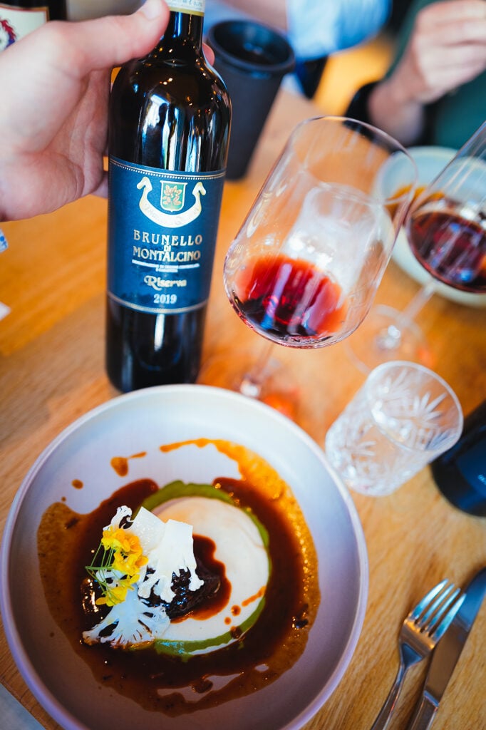 A hand holds a Brunello di Montalcino Riserva 2019 bottle above a plated dish and red wine glass