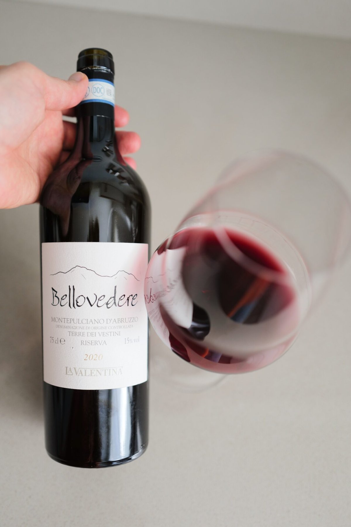 A hand holds the Bellovedere 2020 bottle beside a tilted glass swirling red wine