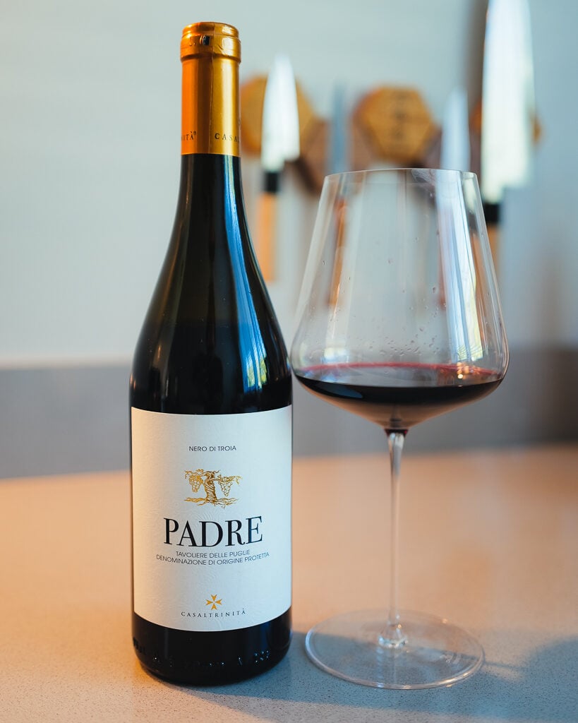 Casaltrinità Padre Nero di Troia red wine bottle with a gold cap beside a glass of red wine