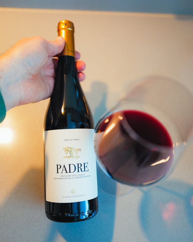 A hand holds the Padre Nero di Troia bottle beside a tilted glass of dark red wine