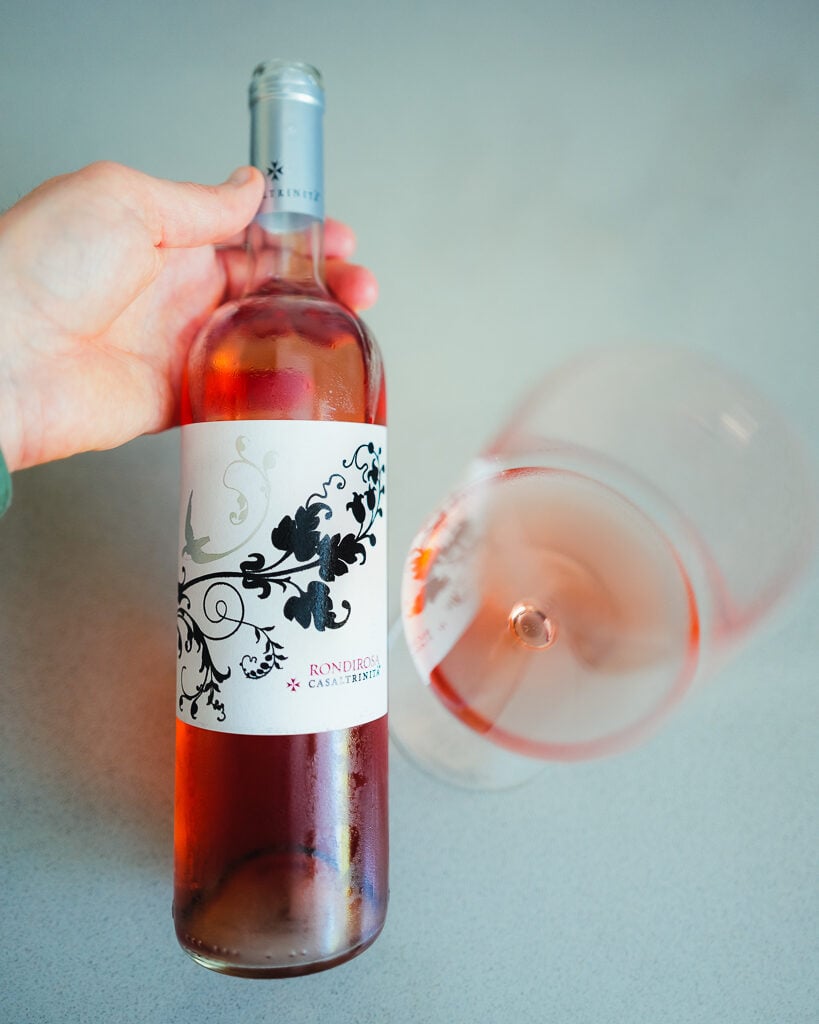 A hand holds the Rondirosa rosé bottle beside a glass of pink wine lying on its side