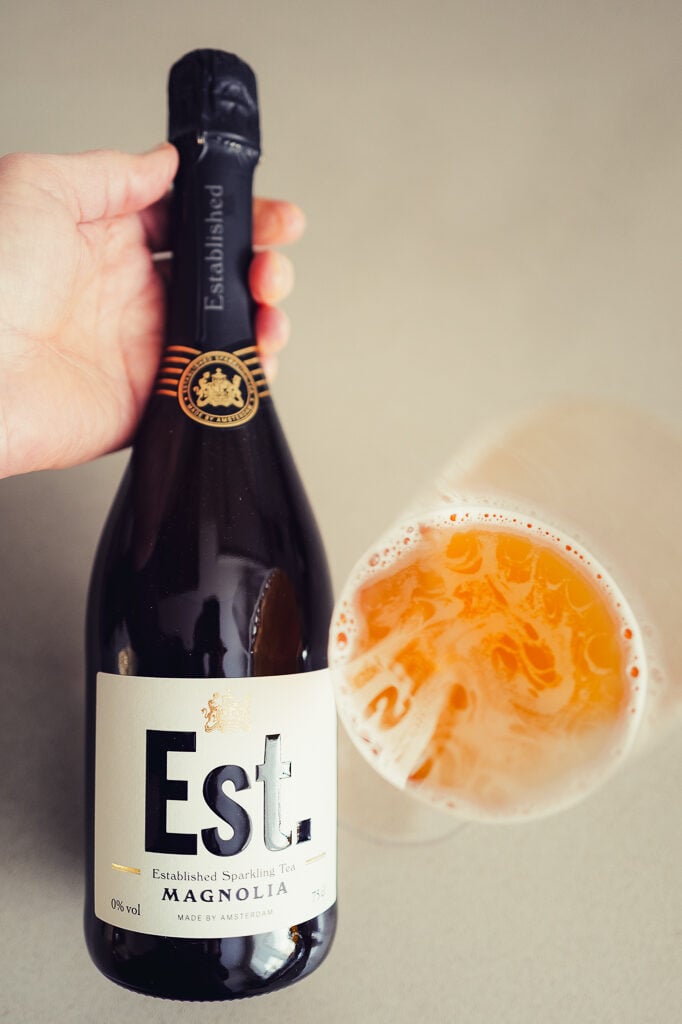 Hand holding the Est. Magnolia bottle above a glass with orange tea bubbles