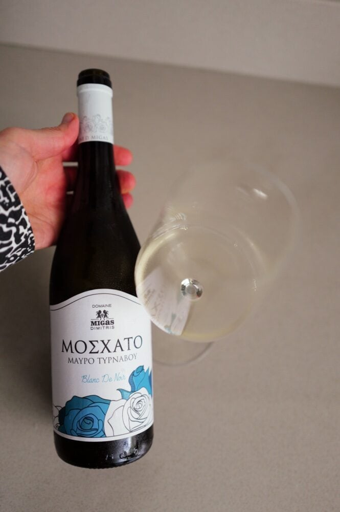 Hand holding the Domaine Migas Moschato Blanc de Noir bottle by a glass of pale white wine