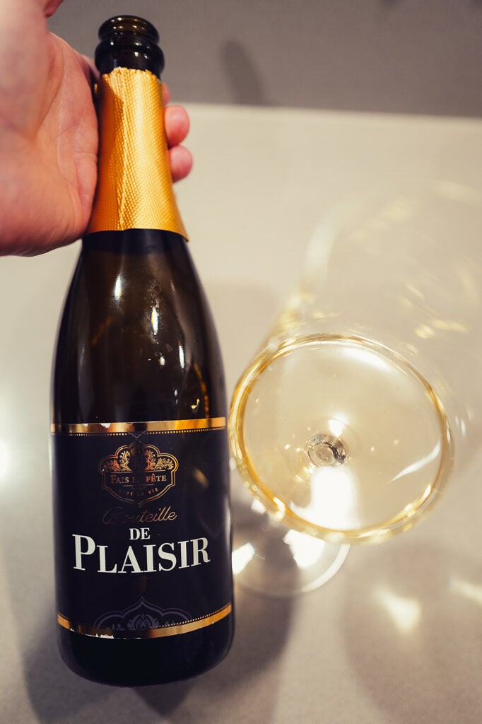 Hand holding the dark Bouteille de Plaisir bottle with gold foil by a glass of golden sekt