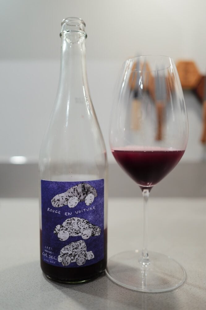 Clear bottle with a purple Rouge en Voiture Cul Sec label beside a glass of dark red drink on a counter