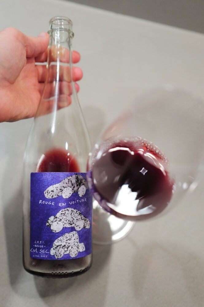 Hand holding the Rouge en Voiture bottle by the neck next to a tilted glass swirling dark red liquid