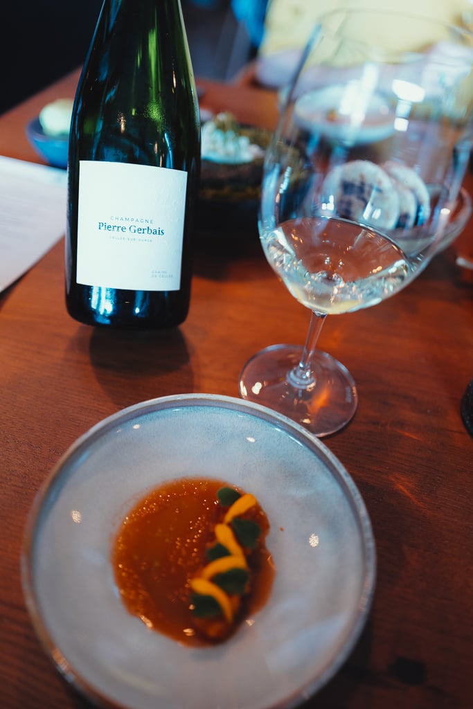A Pierre Gerbais Champagne bottle and a filled glass beside a small course on a plate on a wooden table