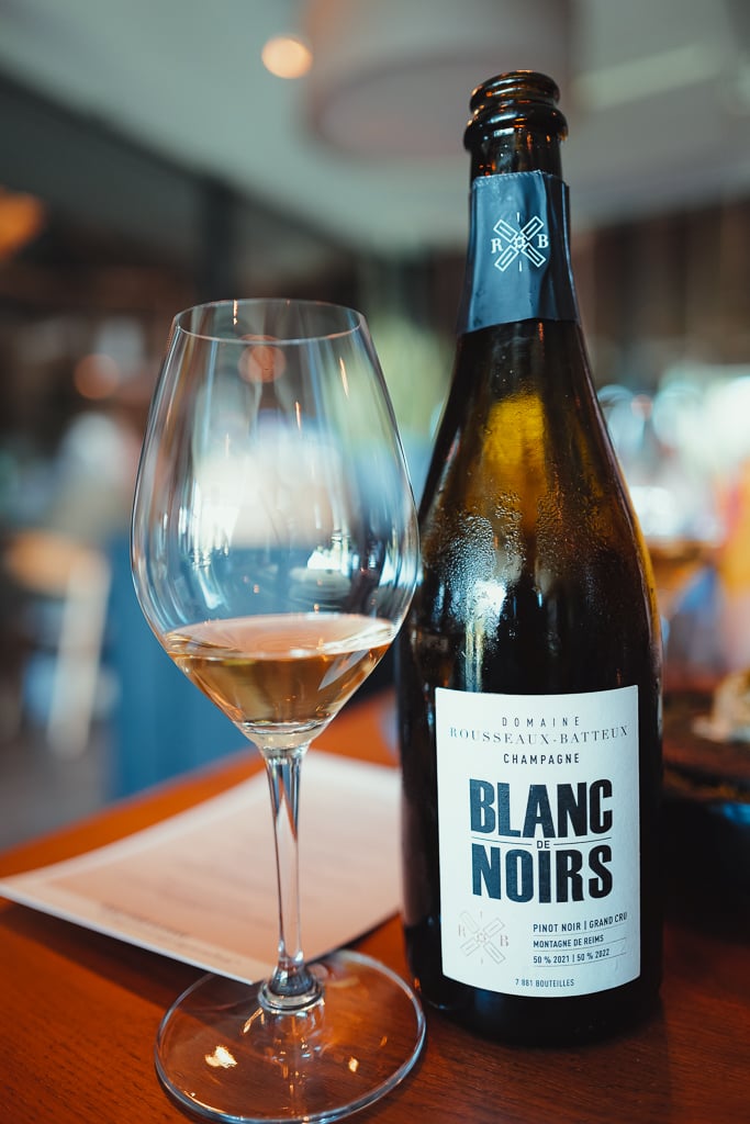 A glass of golden Champagne beside a Rousseaux-Batteux Blanc de Noirs bottle on a wooden table