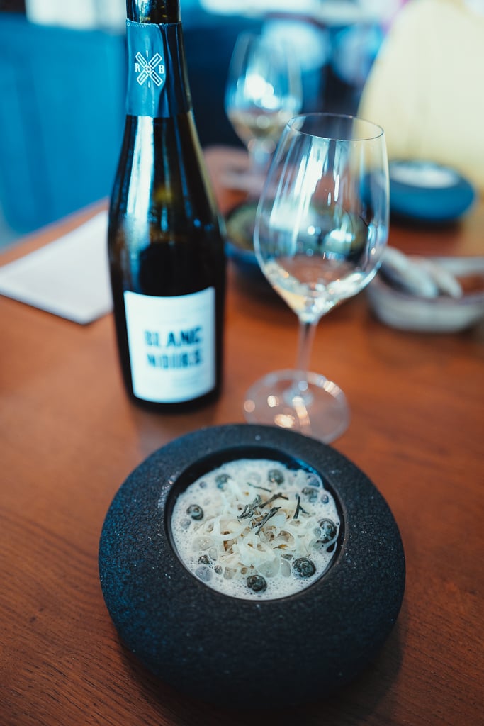 A Blanc Noirs bottle and a glass of Champagne beside a dark stone bowl with a delicate course