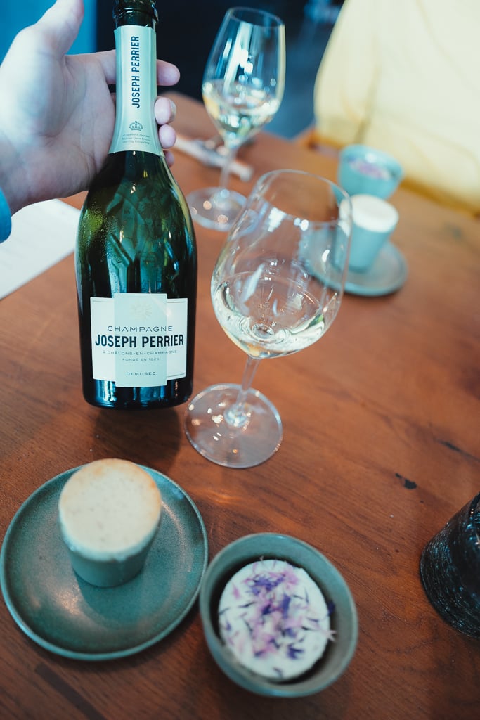A hand holding a Joseph Perrier Demi-Sec bottle by a filled glass and small dessert plates on the table