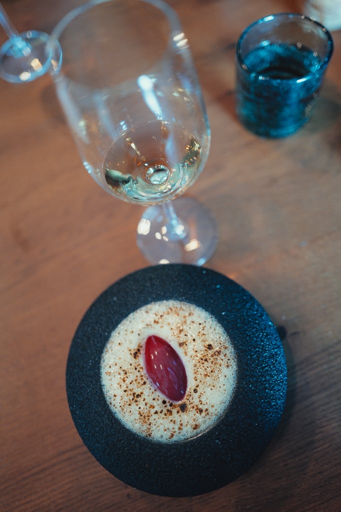 A small dish on a dark stone plate with foam and a single red garnish beside a glass of Champagne