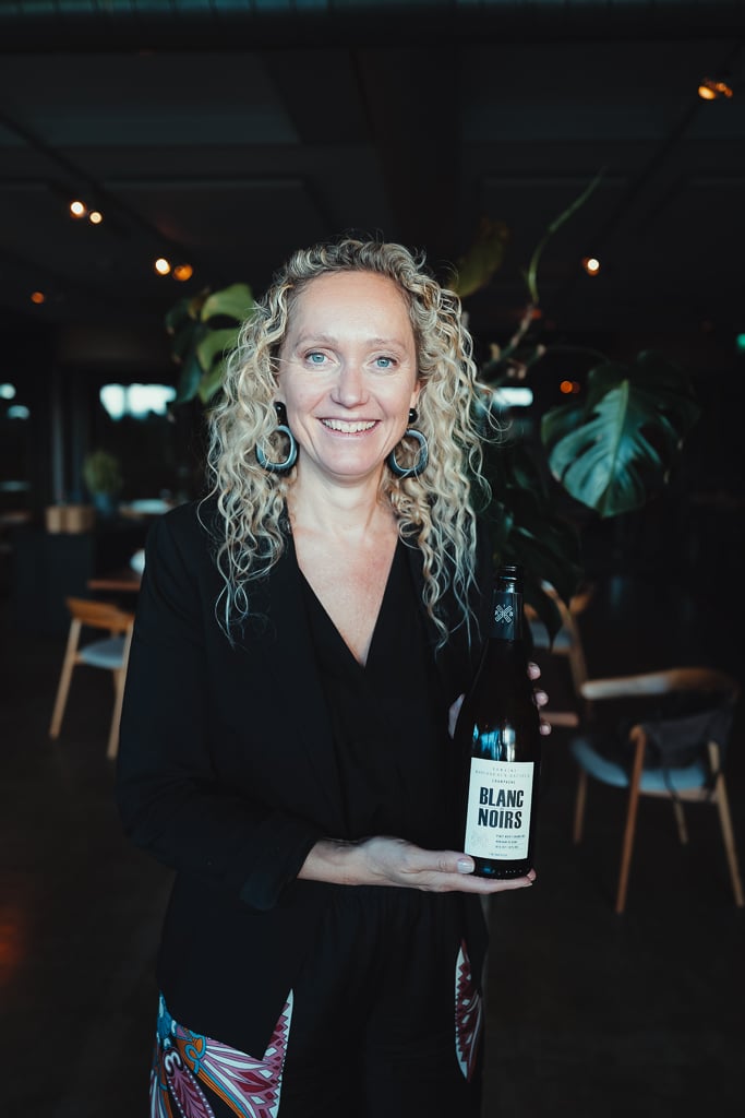 A smiling woman with curly hair holding a Blanc Noirs bottle in the plant-filled restaurant