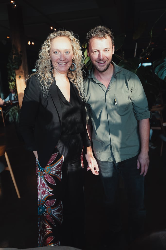 A smiling woman in black and a man in a green shirt standing together in the dim plant-filled restaurant