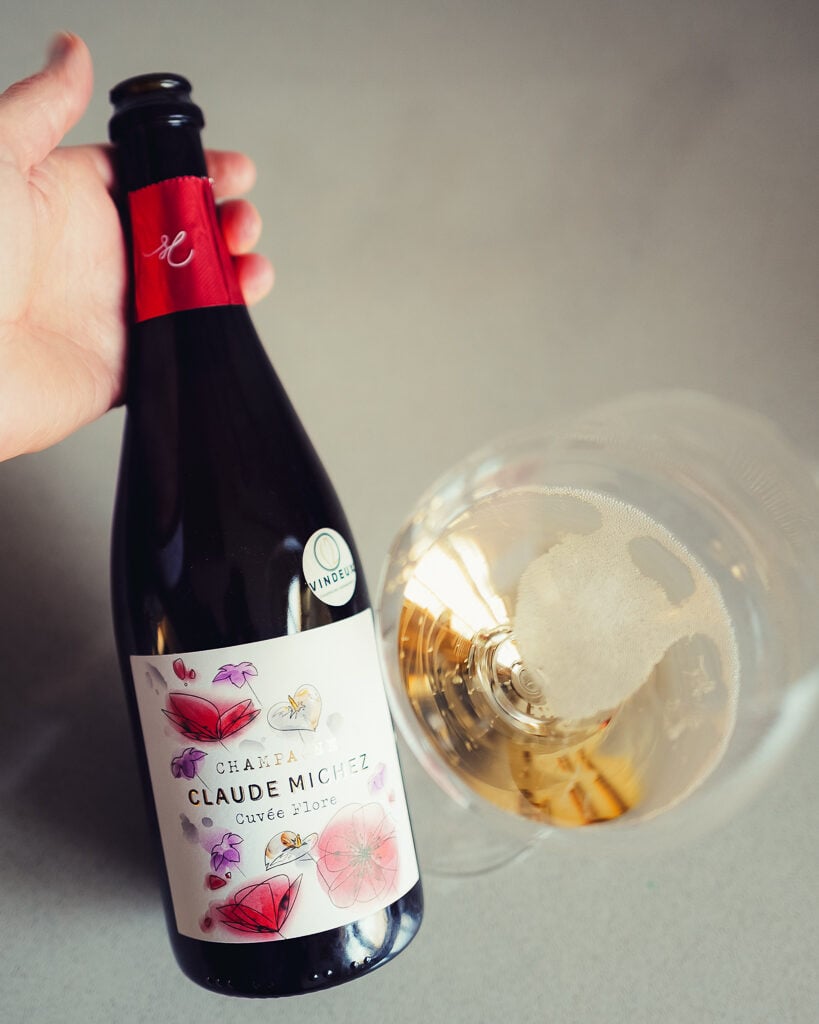 Hand holding the Claude Michez Cuvée Flore bottle beside a tilted glass of golden Champagne