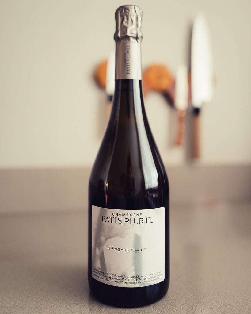 Patis Pluriel Corps Simple Meunier Champagne bottle standing on a counter with knives blurred behind
