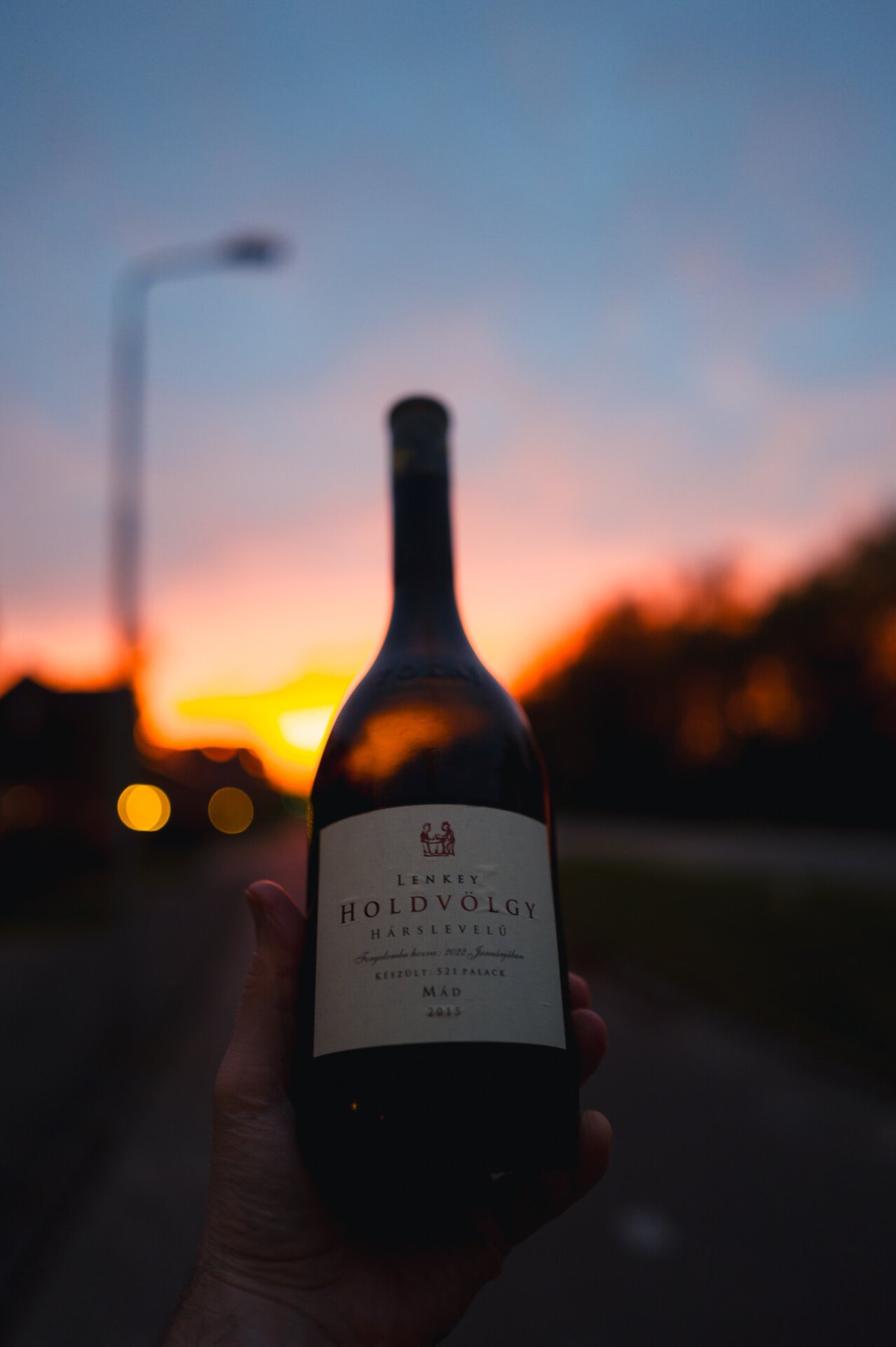 Hand holding the Lenkey Holdvölgy Hárslevelű bottle silhouetted against a glowing orange sunset