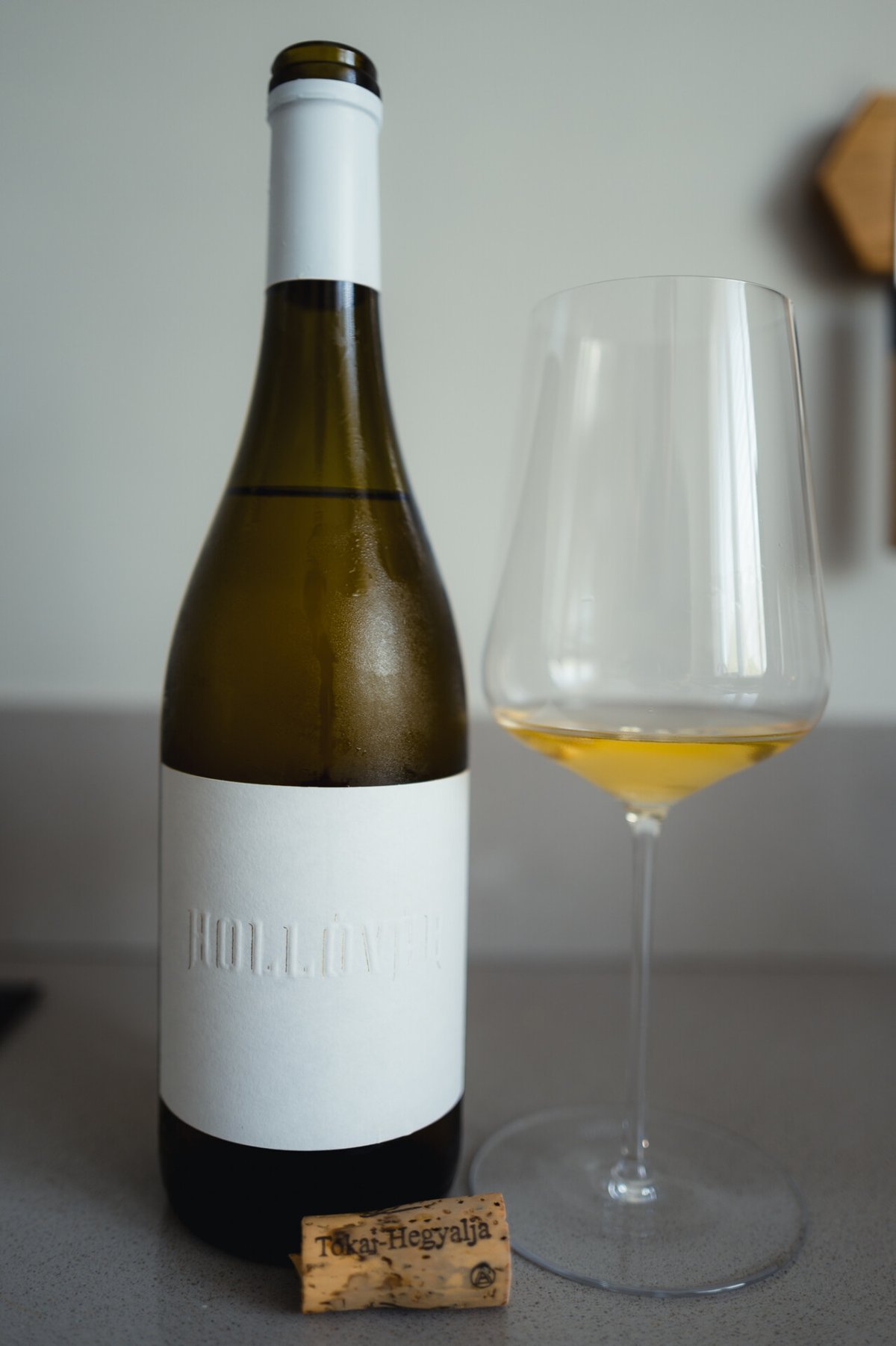 Hollóvár bottle standing beside a glass of golden white wine with a Tokaj-Hegyalja cork on the counter