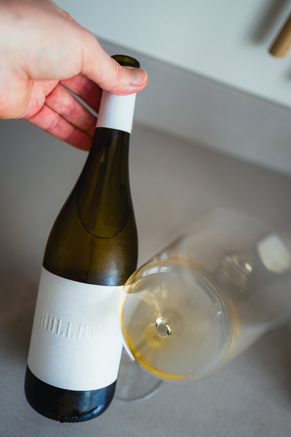 Hand holding the Hollóvár Furmint bottle beside a tilted glass of pale white wine on a counter