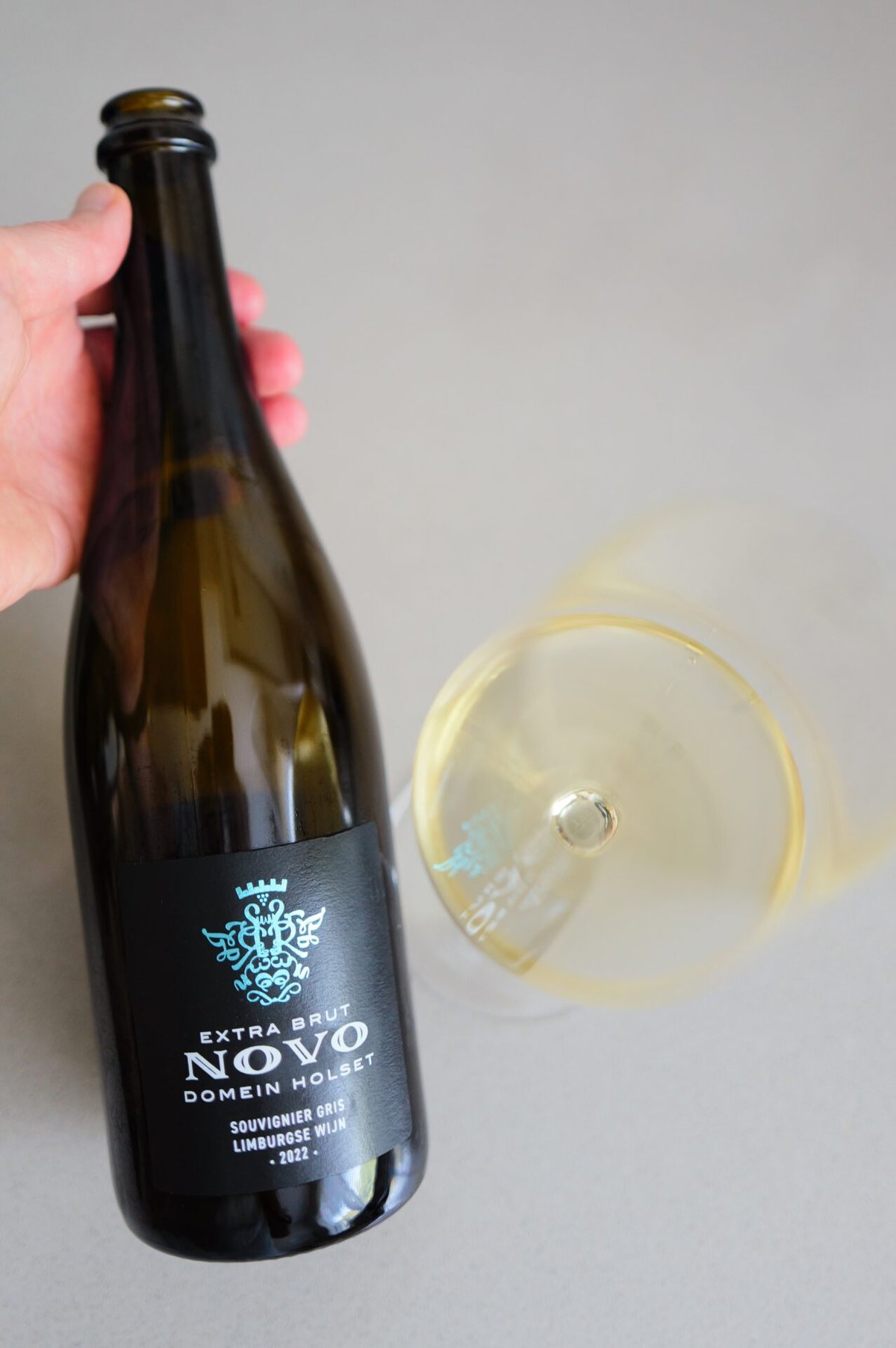 Hand holding the Domein Holset Novo bottle beside a tilted glass of golden sparkling wine
