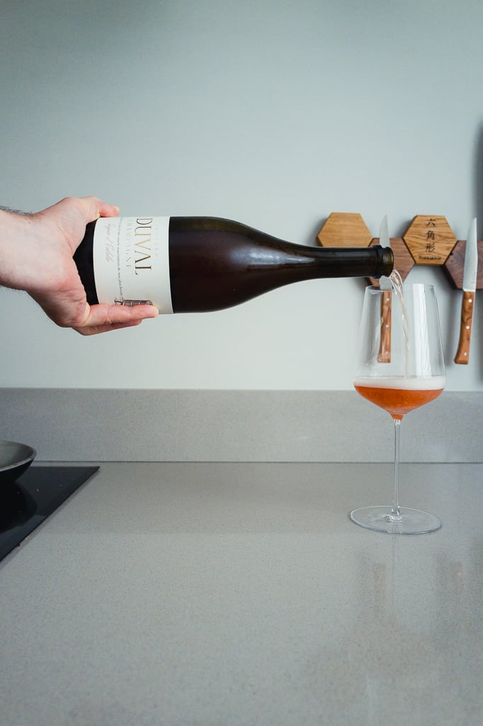 Hand pouring sparkling rosé from an Edouard Duval bottle into a stemmed glass on a kitchen counter