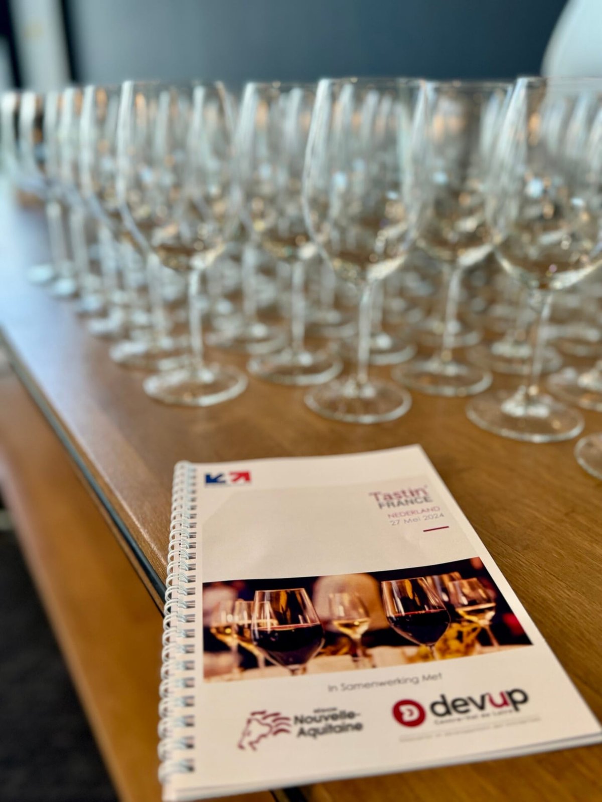 Rows of empty wine glasses on a wooden table behind a Tastin' France 2024 Nederland tasting booklet