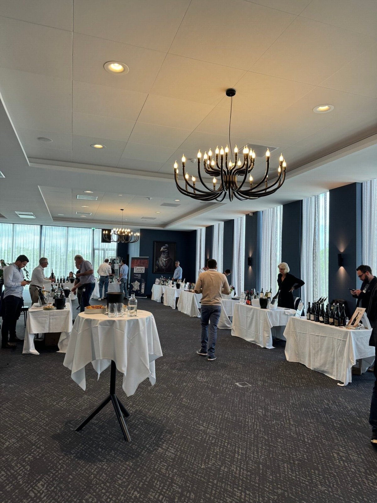 Tasting room with white-clothed high tables, wine bottles and visitors under a black chandelier