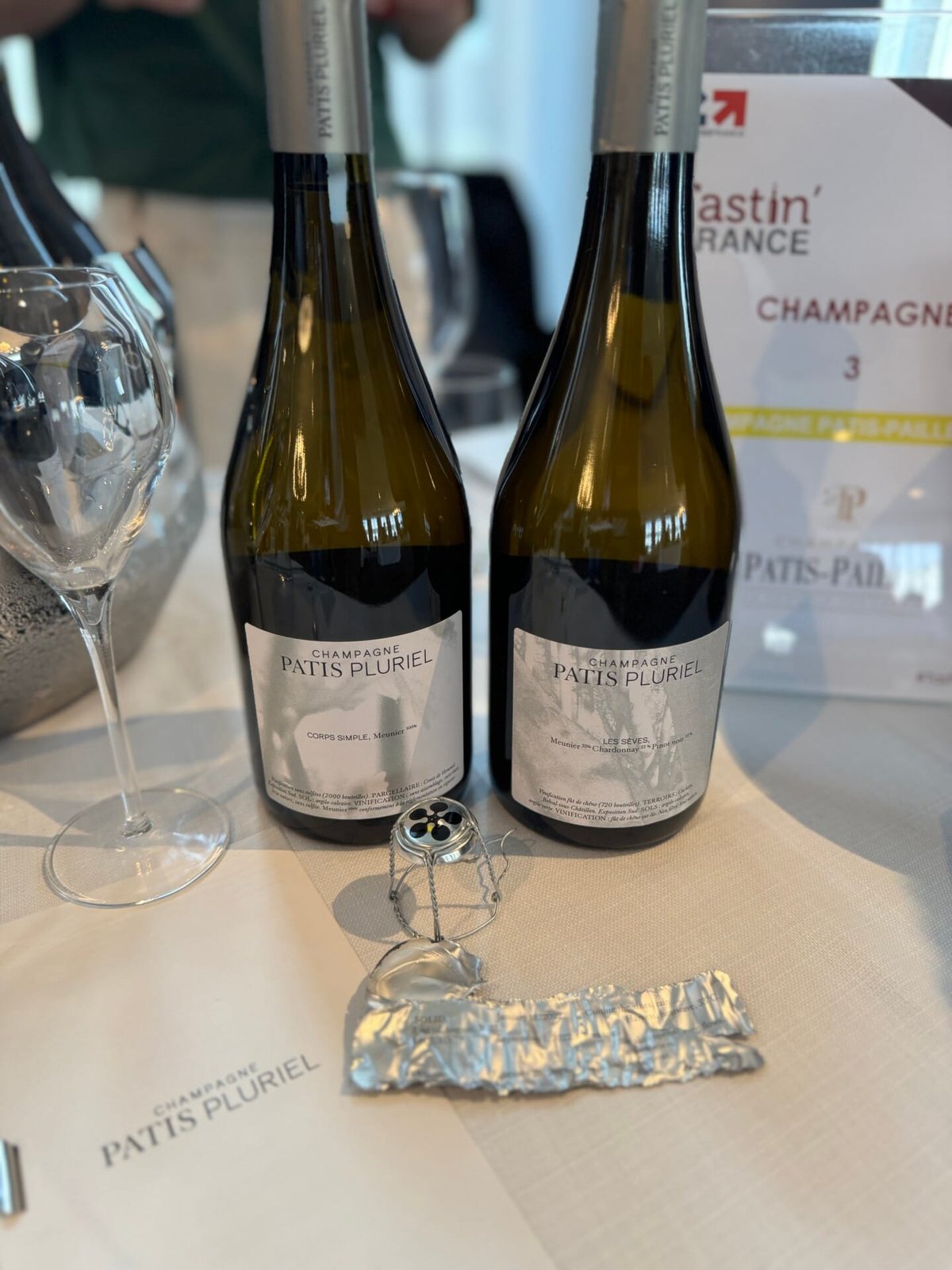 Two Champagne Patis Pluriel bottles on a table with an opened wire cage and foil capsule