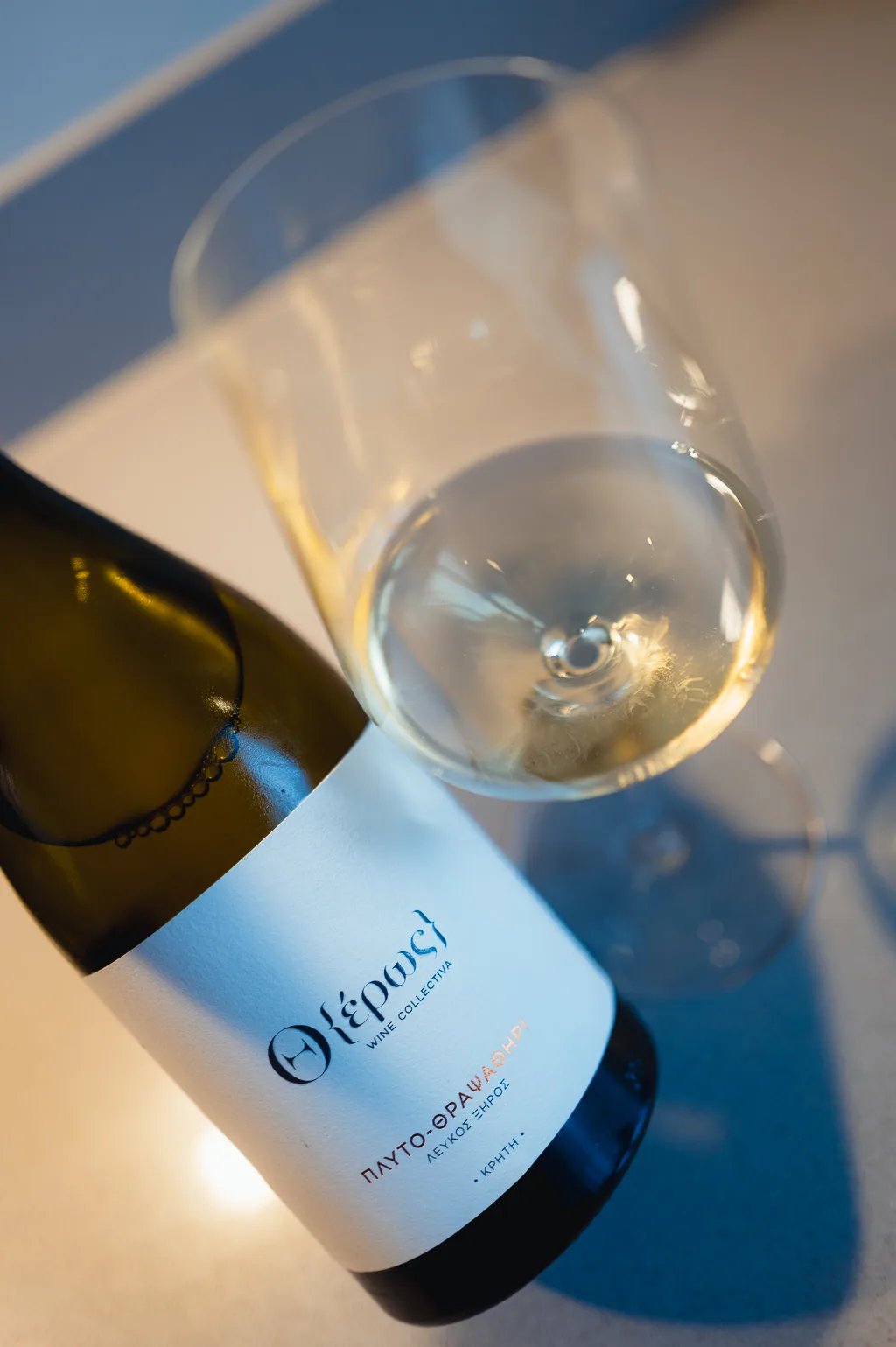 Reclining Theros Wine Collectiva Plyto-Thrapsathiri bottle next to a tilted glass of white wine