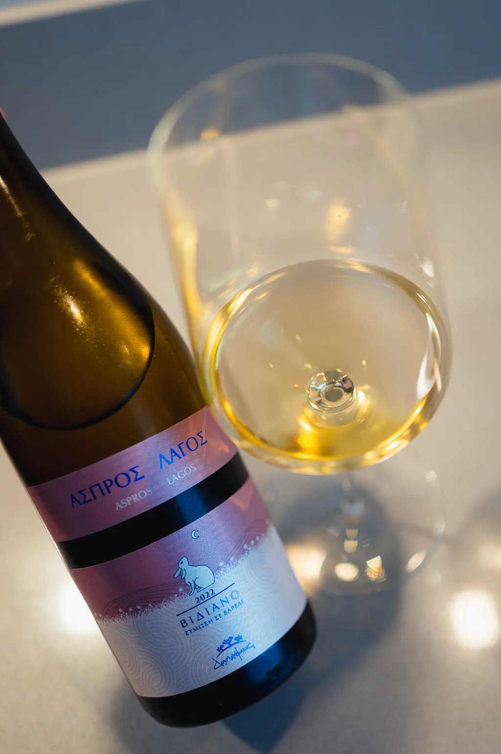 Reclining Douloufakis Aspros Lagos bottle next to a tilted glass of pale white wine