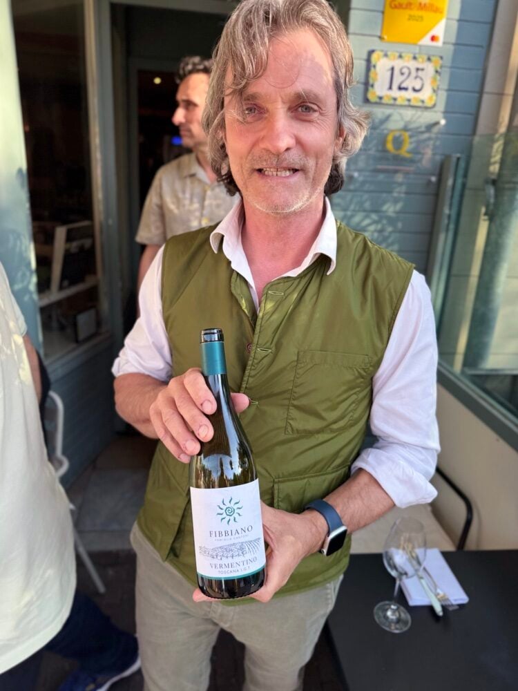 Man in a green vest holding a bottle of Fibbiano Vermentino Toscana white outside a wine bar