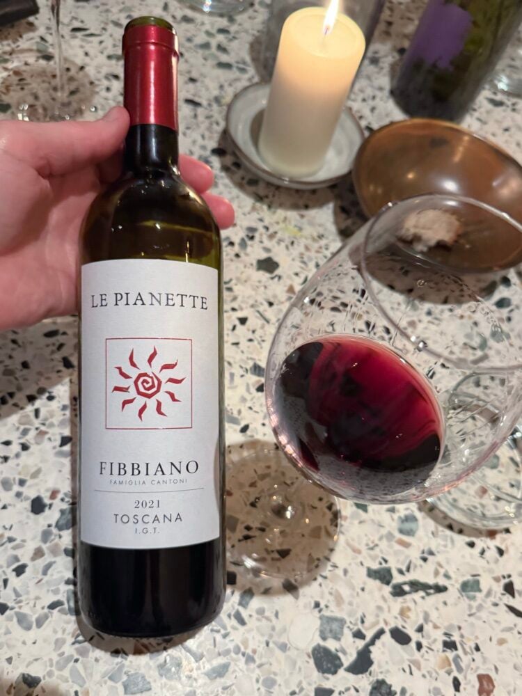 Hand holding a bottle of Fibbiano Le Pianette Toscana 2021 red next to a glass of red wine