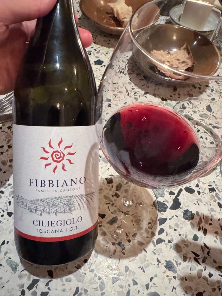 Hand holding a bottle of Fibbiano Ciliegiolo Toscana red beside a glass of red wine and bread
