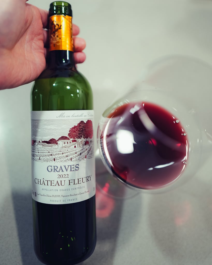Hand holding a bottle of Graves 2022 Château Fleury red next to a glass of red wine
