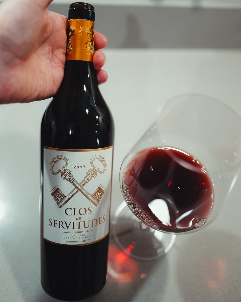 Hand holding a bottle of Clos des Servitudes 2017 Cadillac red beside a glass of red wine