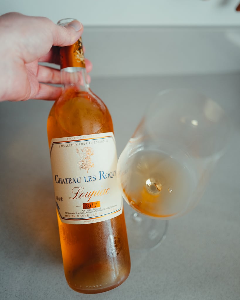 Hand holding a bottle of Château Les Roques 2017 Loupiac sweet white beside a tilted glass