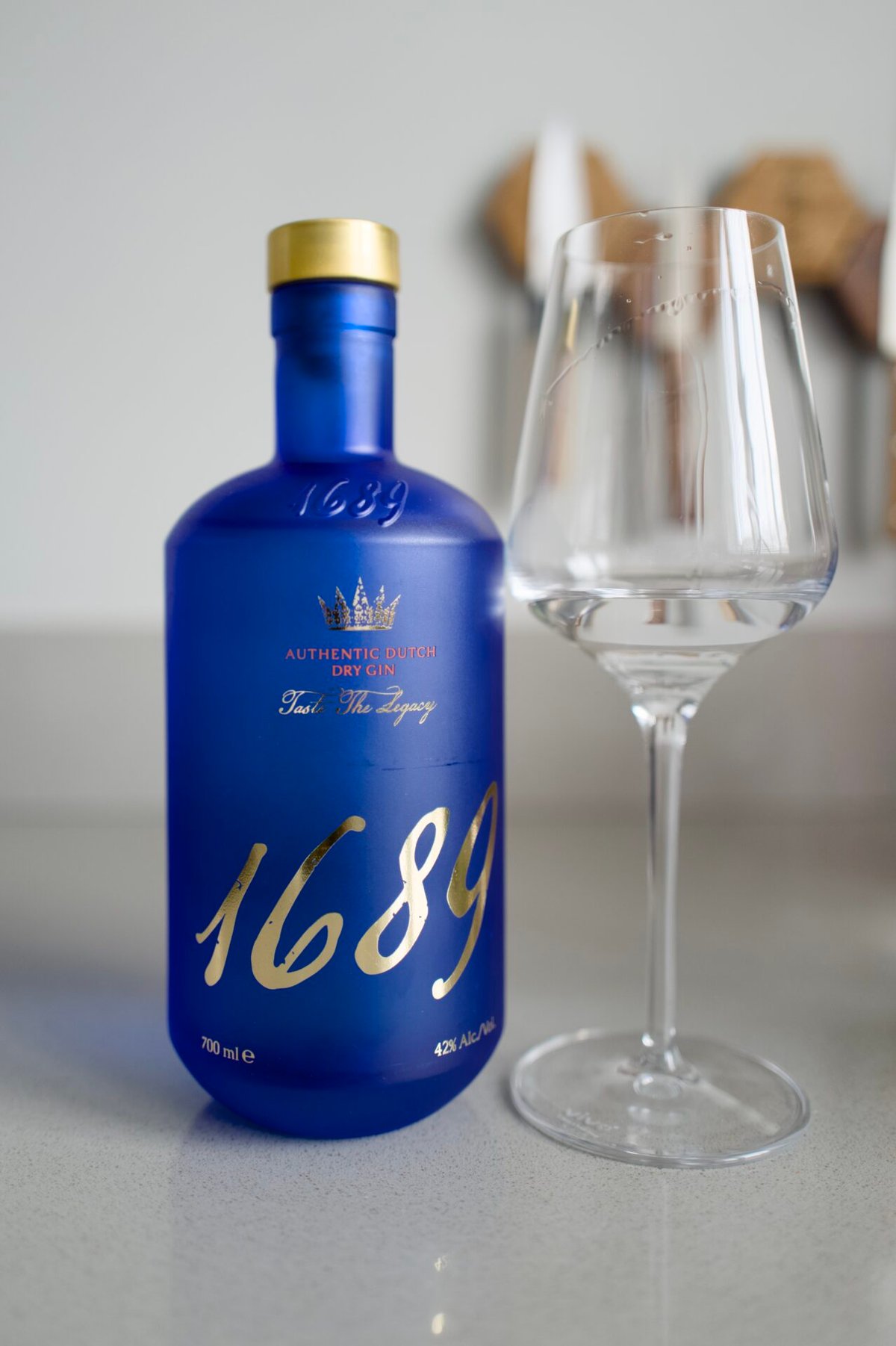 Blue frosted Gin 1689 Dutch Dry bottle with gold cap and lettering beside an empty stemmed glass
