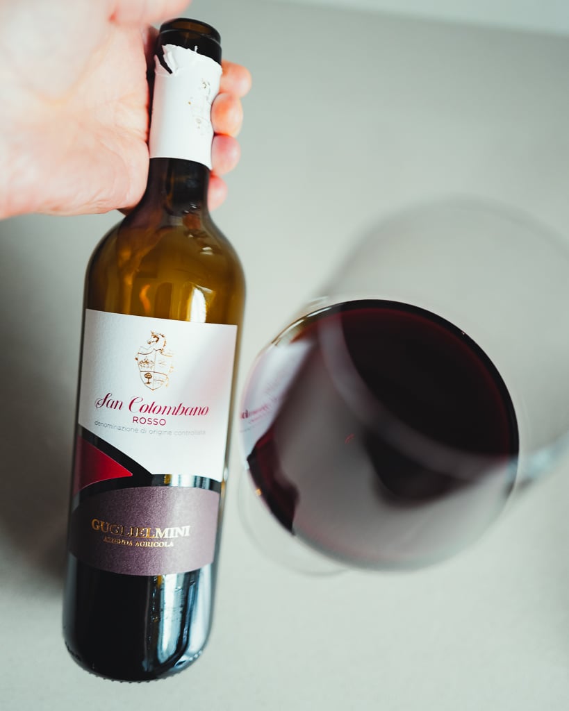 Hand holding the Guglielmini San Colombano Rosso bottle over a glass of red wine seen from above