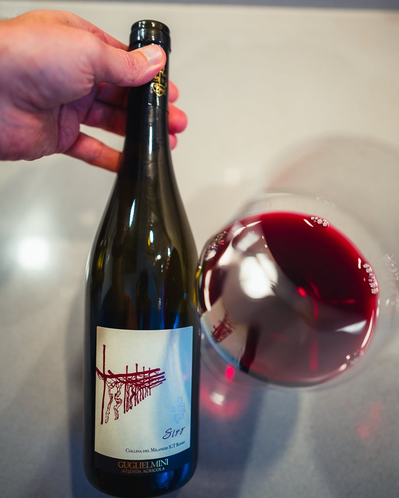 Hand holding the Guglielmini Siro bottle with vine-sketch label over a glass of red wine
