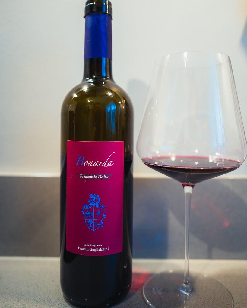 Guglielmini Bonarda Frizzante Dolce bottle with magenta label beside a glass of red wine