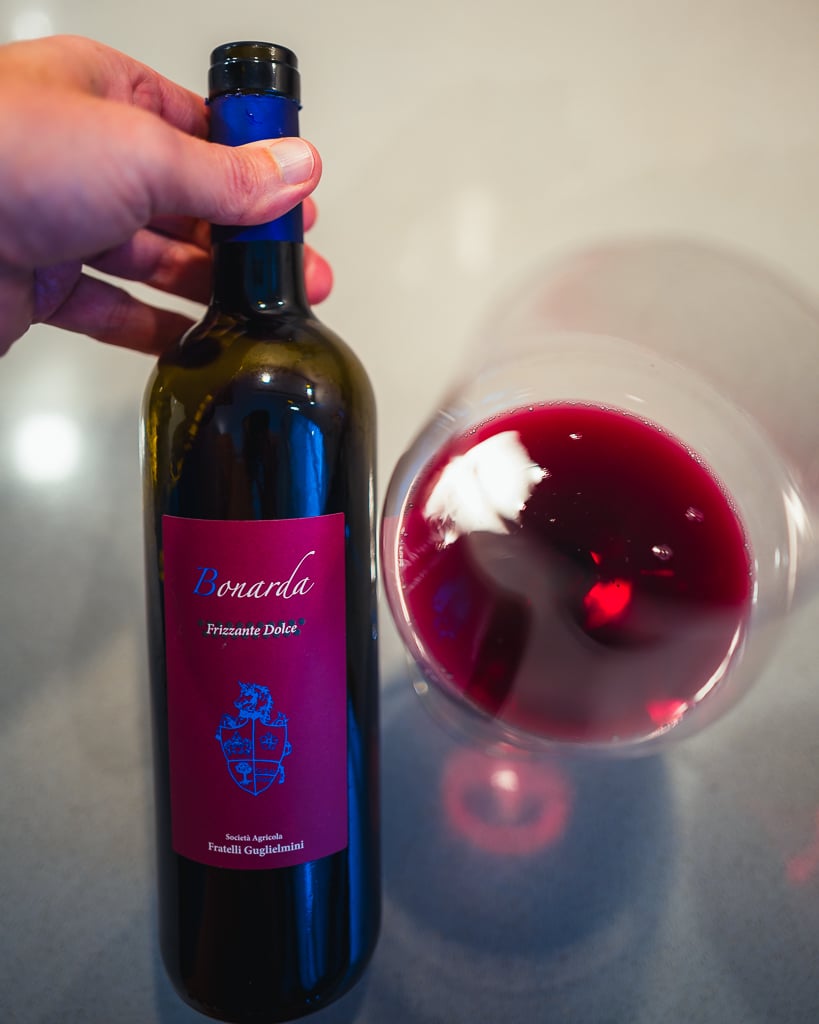 Hand holding the Guglielmini Bonarda Frizzante Dolce bottle over a glass of bright red wine