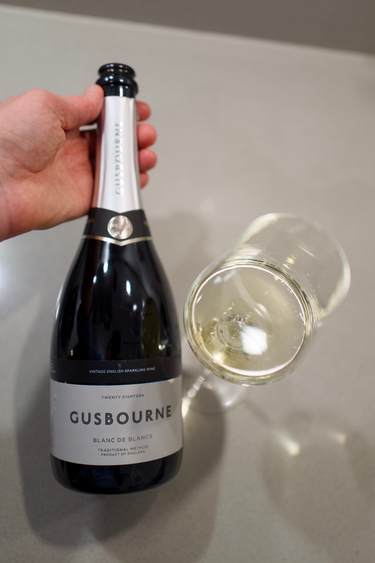 Hand holding a Gusbourne Blanc de Blancs bottle next to a glass of pale sparkling wine