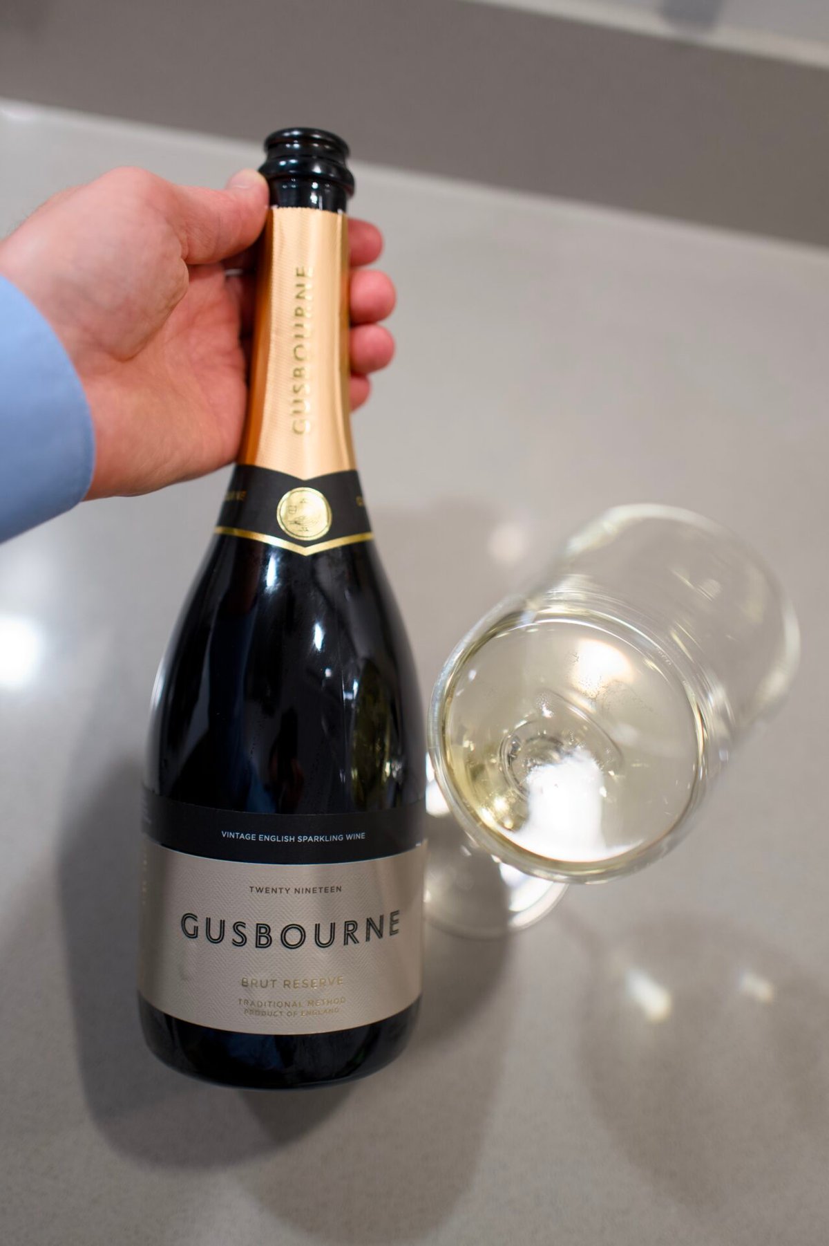 Hand holding a Gusbourne Brut Reserve bottle beside an empty sparkling wine glass