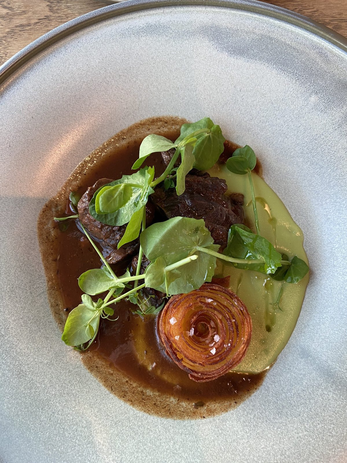 Braised meat in dark jus with pea shoots and a crisp onion ring in a grey bowl