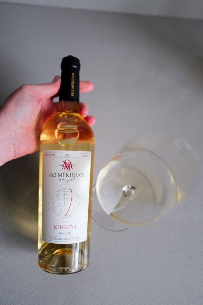 Hand holding the 45.3 Meridian Khikhvi 2024 bottle above a glass of pale white wine