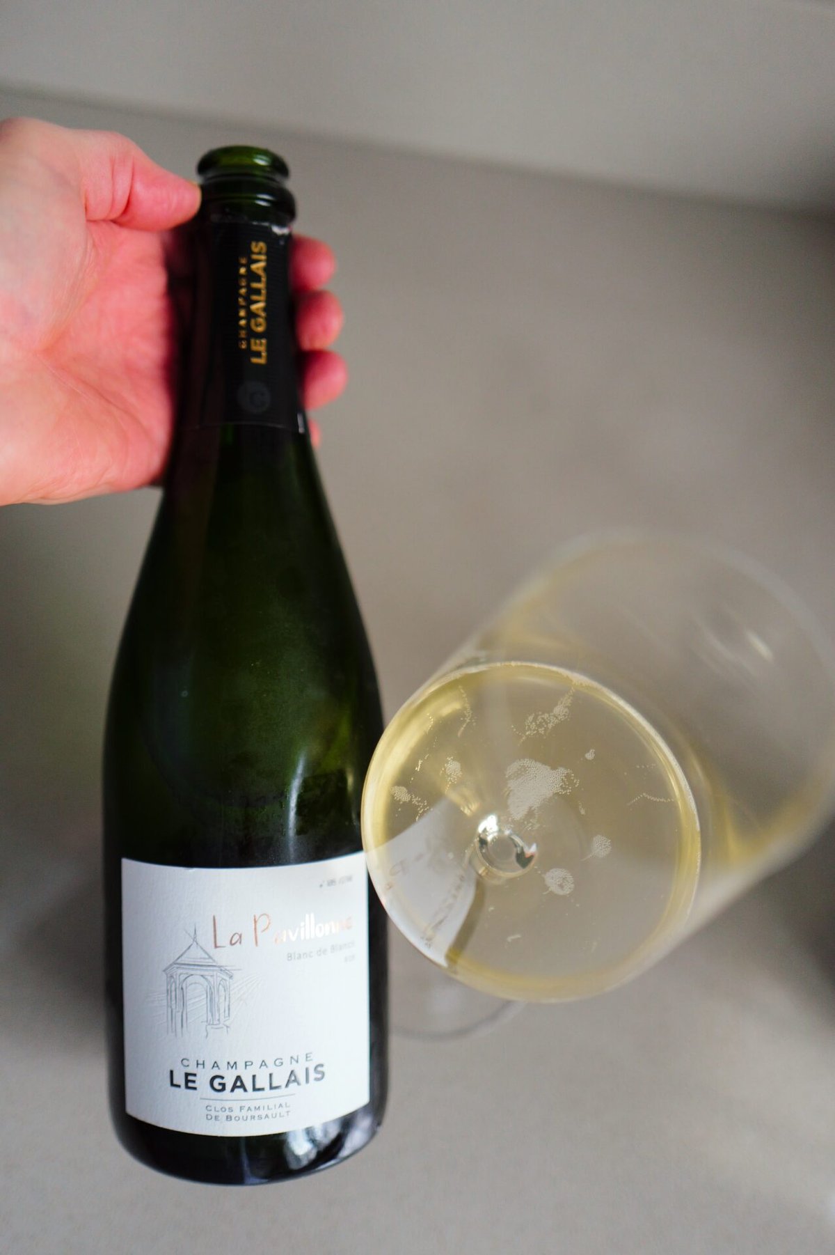 Hand holding the La Pavillonne Le Gallais bottle beside a tilted glass of golden champagne