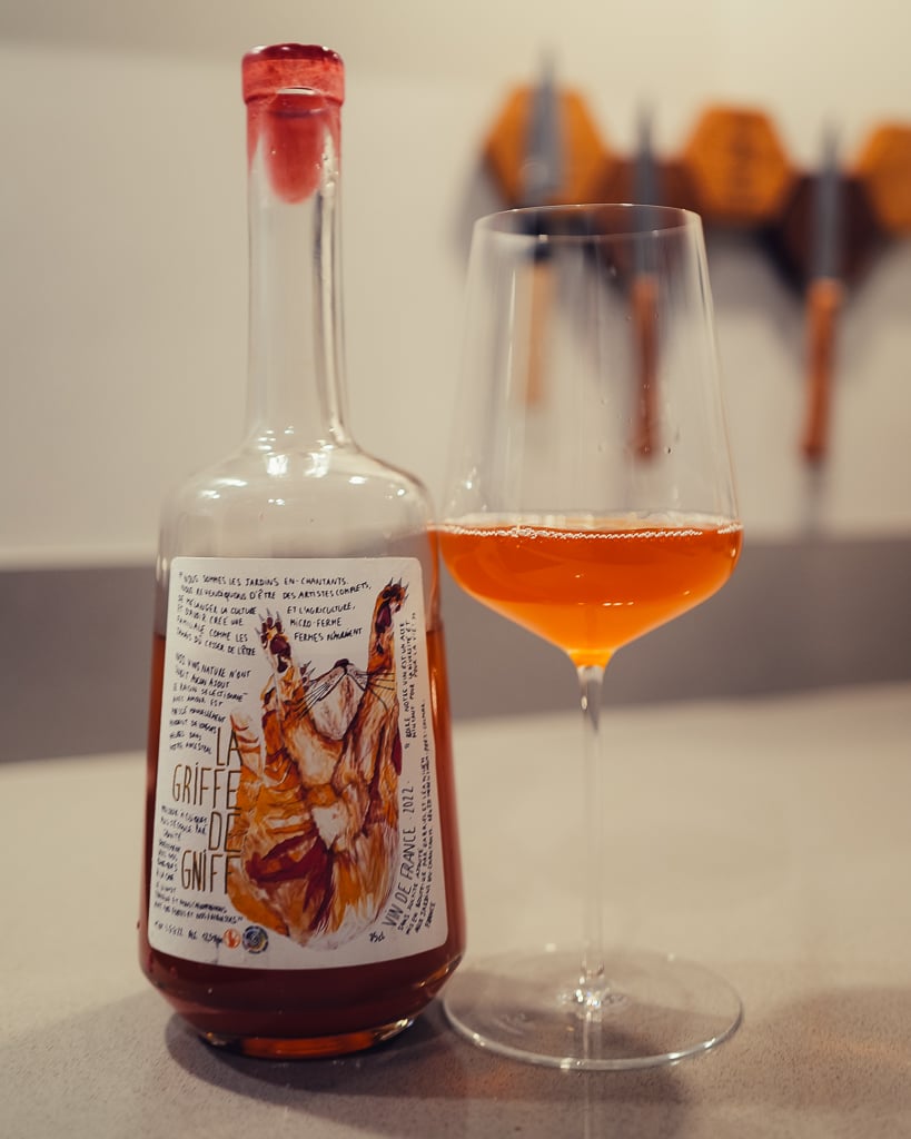 Hand holding the La Griffe de Gniff orange wine bottle beside a glass of amber wine