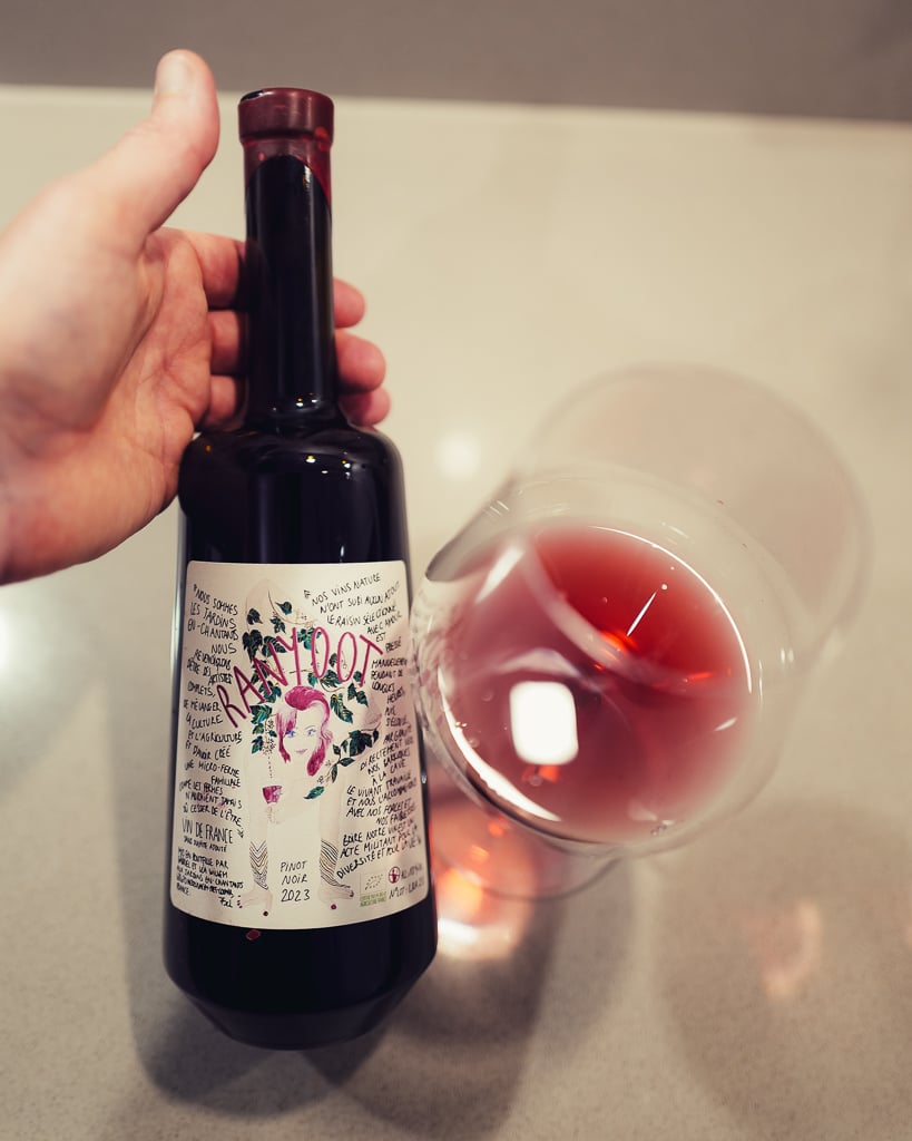 Hand holding the Ranyoot 2023 Pinot Noir bottle with its illustrated label beside a glass of red wine